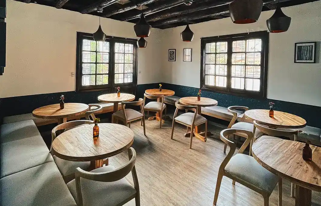Interior de un restaurante con mesas redondas de madera, sillas y bancas bajo vigas de madera oscura